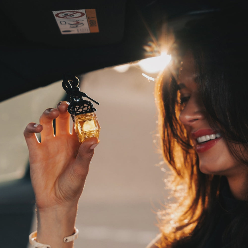 Een vrouw houdt in de auto een Oduree-autoparfumhanger omhoog in warm tegenlicht.